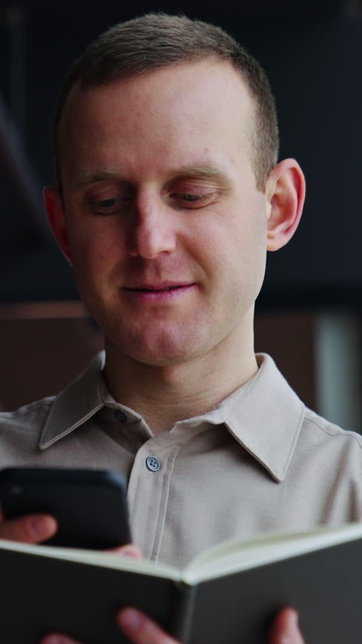 Mature man holds the notebook opened in front of him and print something on the phone. He throws interested look into the window. Vertical video