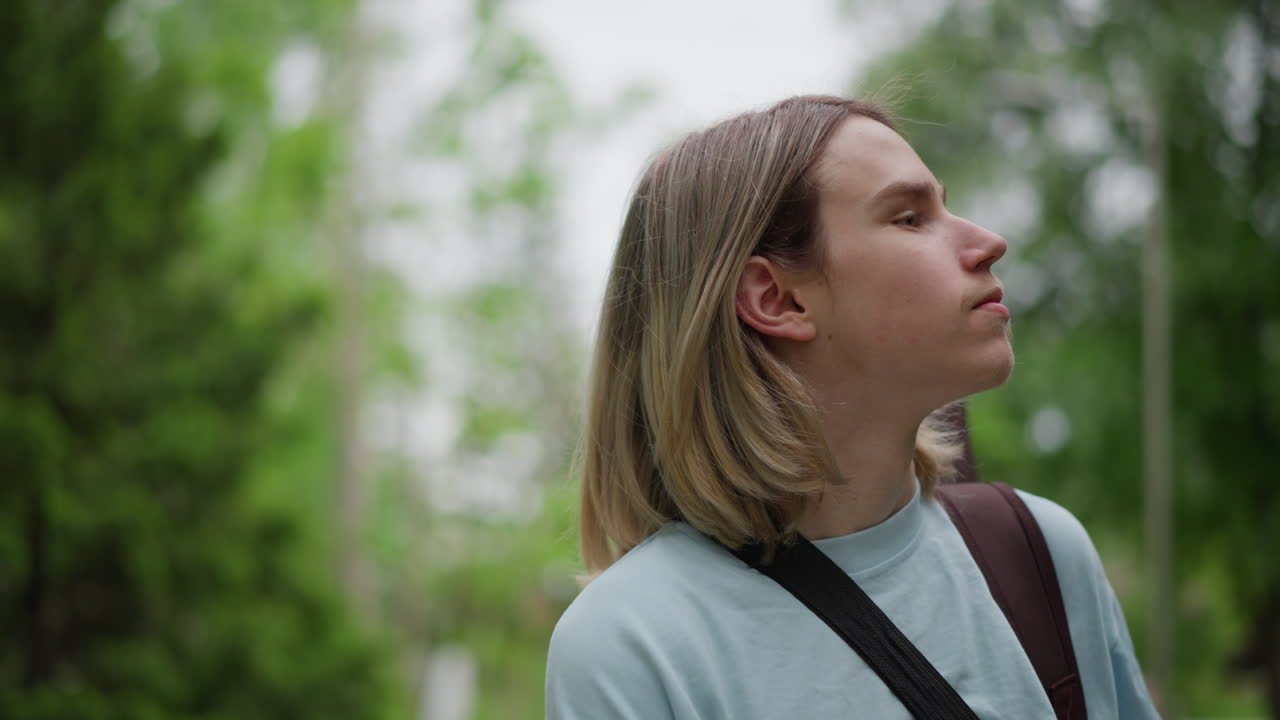 jovem guitarrista branco ao ar livre, carregando um estojo de guitarra nas costas, andando por um parque arborizado, expressão contemplativa, clima de compositor indie, luz suave e nublada, suéter casual, alça de ombro