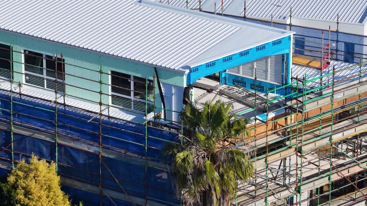 Vista aerea di una casa contemporanea in costruzione, circondata da impalcature e palme, ripresa in pieno giorno con un'angolazione della telecamera statica.