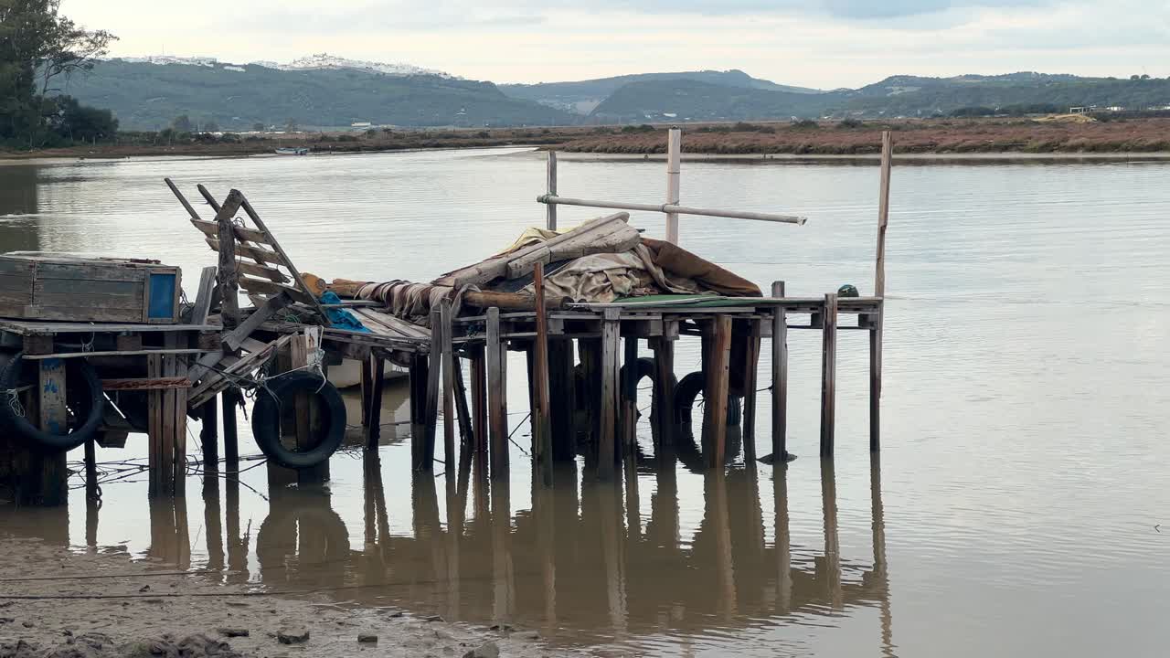 Inquadratura statica di un molo rustico in legno con pneumatici appesi usati come parabordi per barche su acque calme dell'estuario vicino a Barbate Cadice