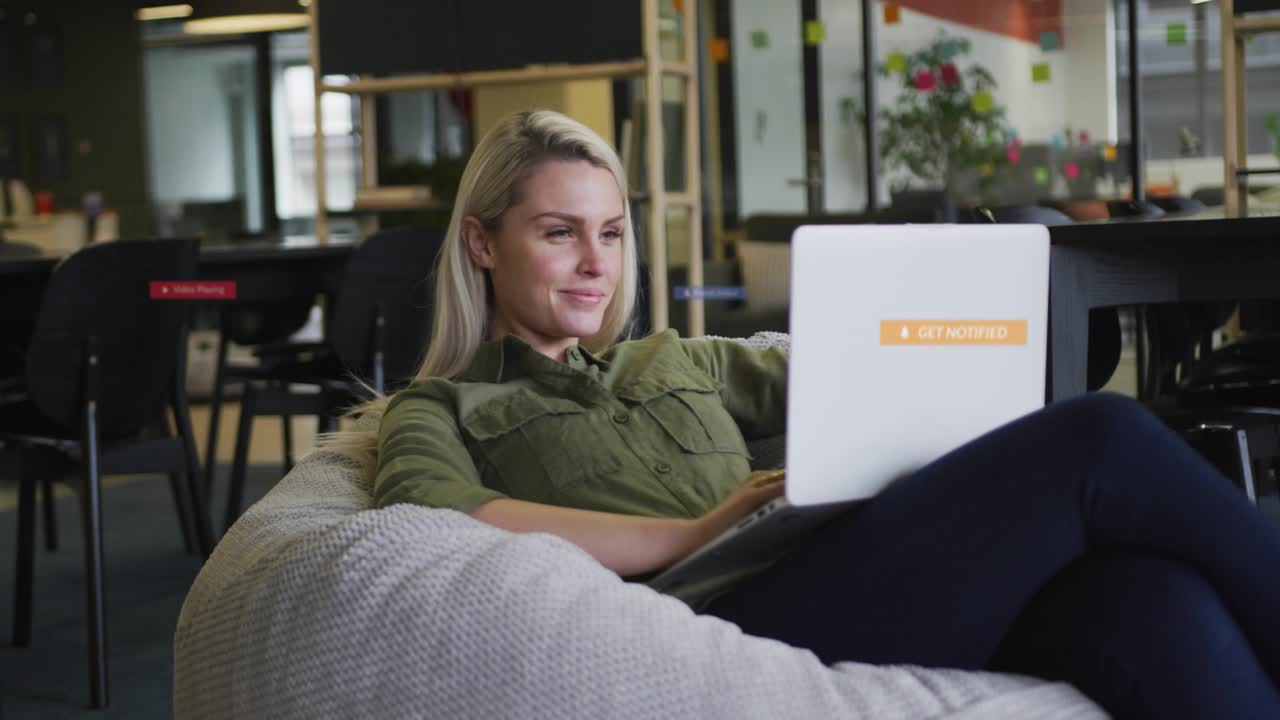 Woman sitting on beanbag in office typing on laptop starting animated alerts over lap for marketing