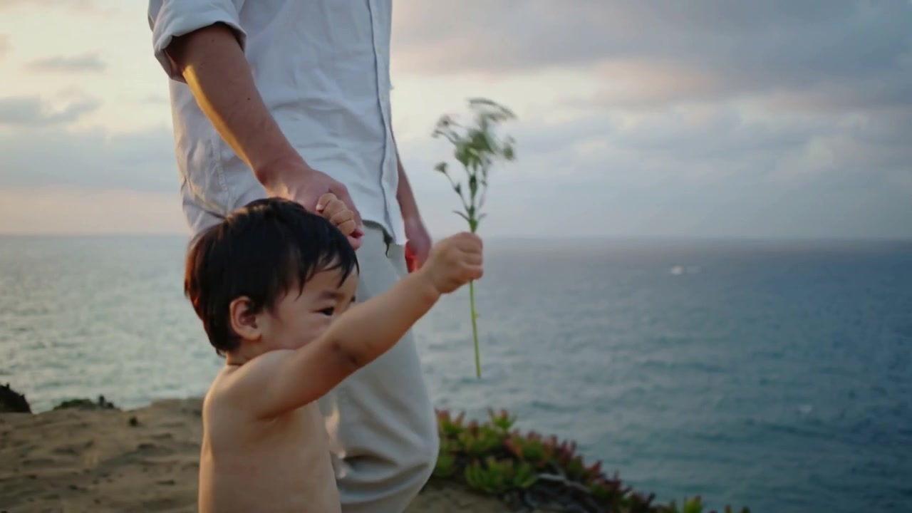 崖の上で夕日を浴びながら白い花を振って歩く幼児、風が波を誘い、大人がシャツの中で手を握っている