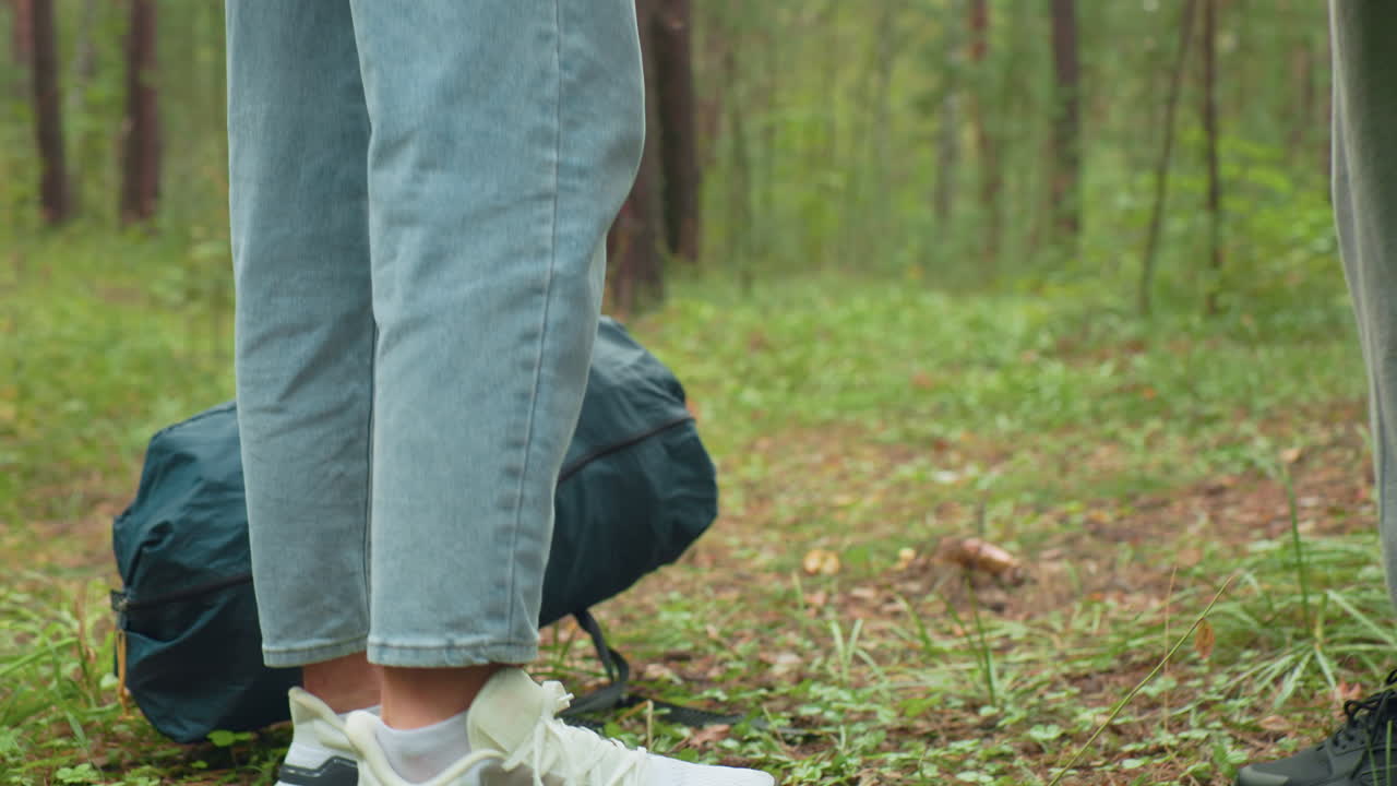 Rear view of young couple pausing in woodland to drop camping bags, showing close-up of green duffel and maroon backpack while surrounded by lush greenery and natural forest background