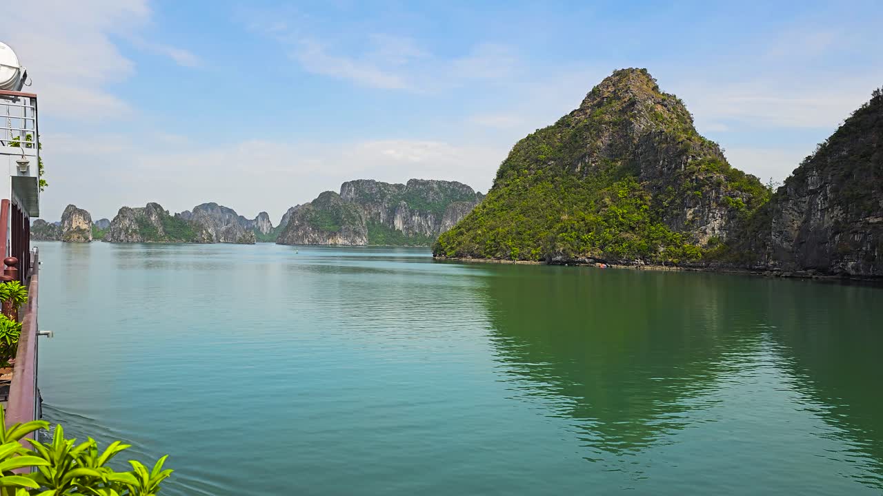 A mesmerizing view from a cruise ship's deck in Ha Long Bay captures emerald green waters surrounding towering limestone karsts, steep cliffs under a serene blue sky