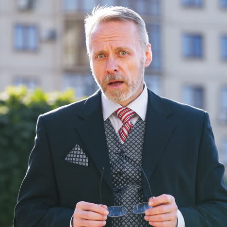 Portrait of mature businessman holding eyeglasses outdoors. Middle aged man with grey hair wearing elegant suit and glasses standing in the street and telling something happily.