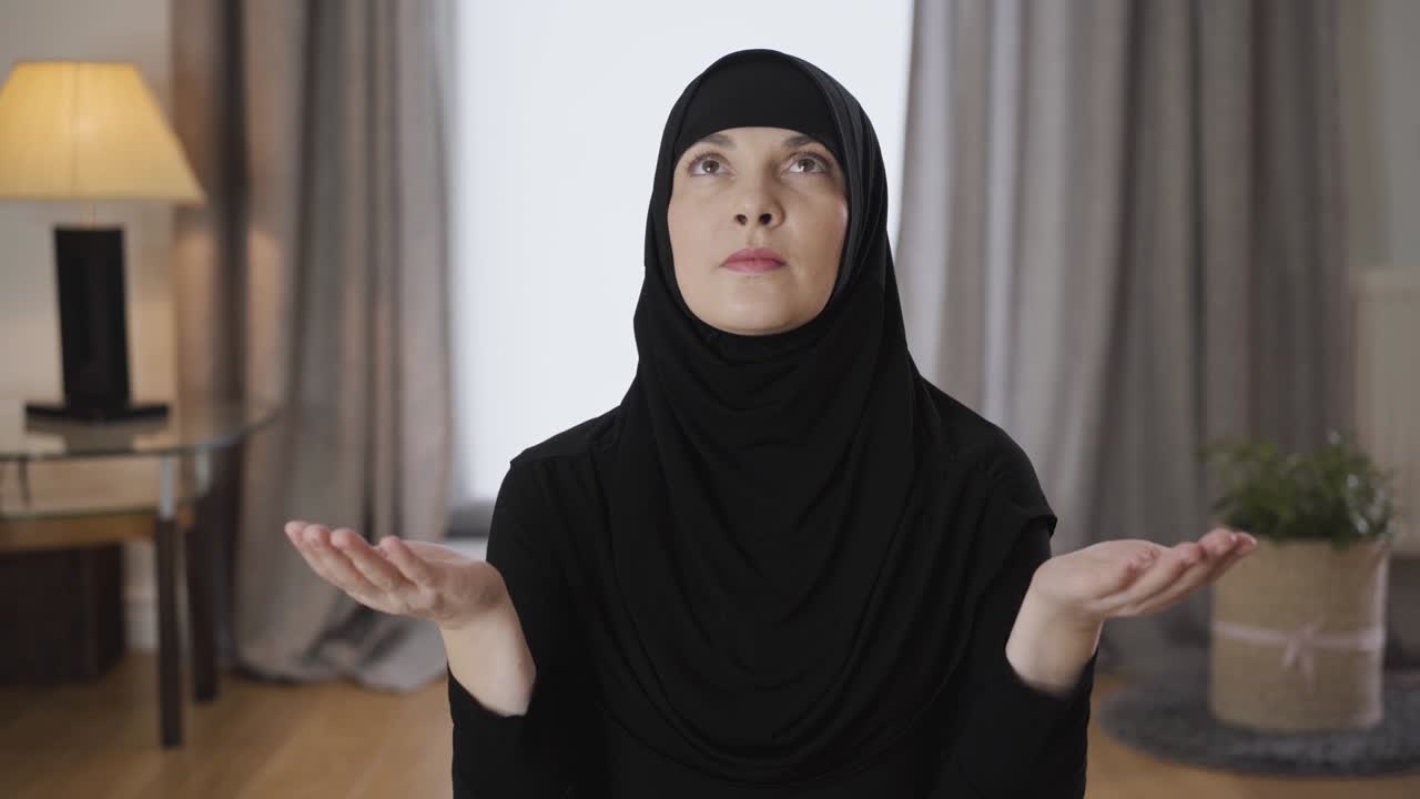 Portrait of beautiful Muslim woman sitting with raised hands and looking up. Young lady in black hijab praying at home. Religion, traditional culture.