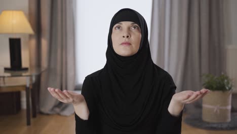 portrait of beautiful muslim woman sitting with raised hands and looking up. young lady in black hijab praying at home. religion, traditional culture.