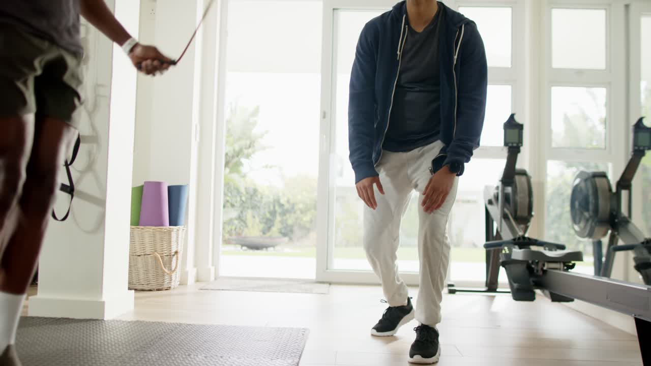 Male coach in home gym seeing trainee skipping rope, crouching, clapping and guiding form near mat