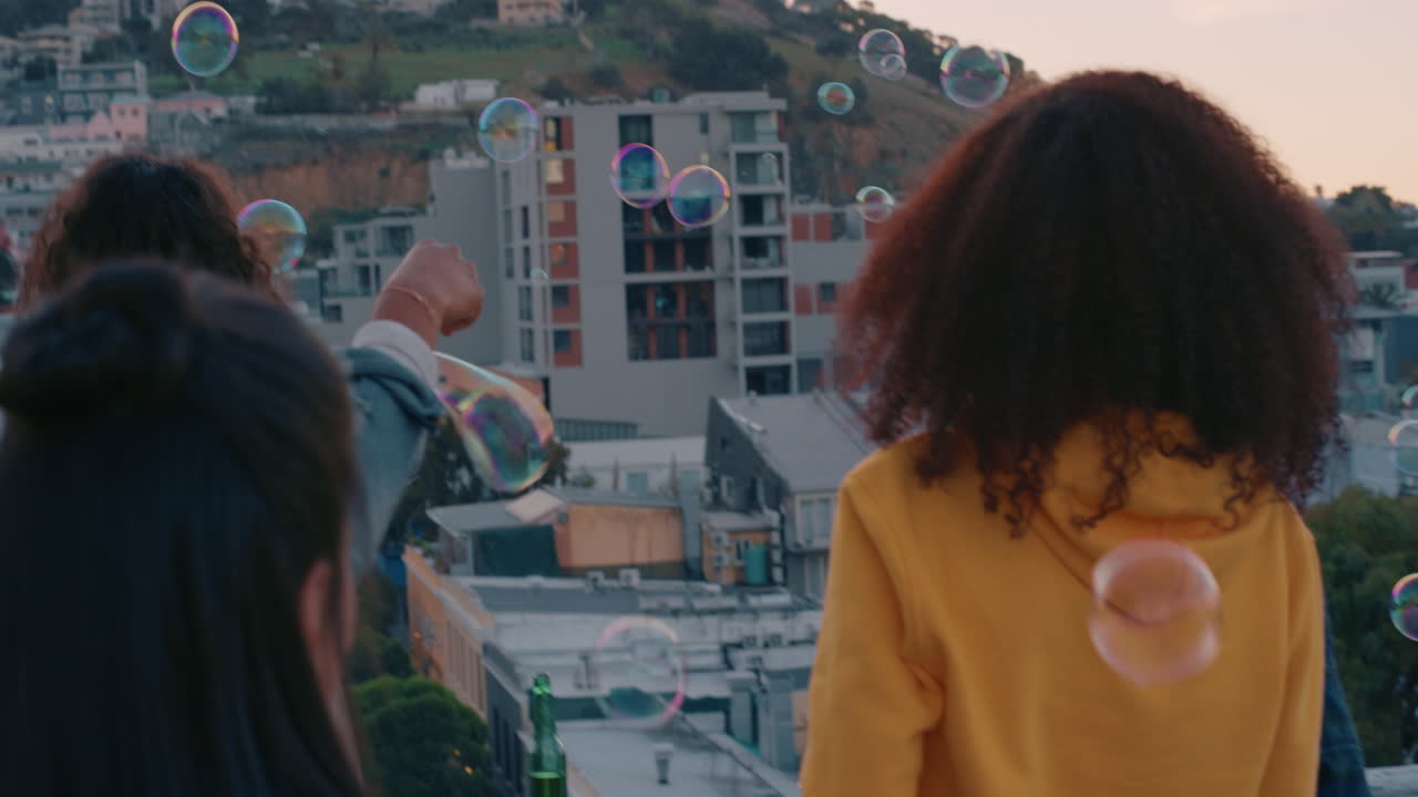 해가 지는 날 지붕에서 거품을 불어넣는 행복한 친구 그룹이 배경으로 도시 스카이라인과 함께 무의식적인 주말을 즐기고 있습니다.