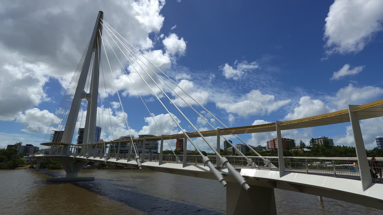 New Kangaroo Point Green Bridge over the Brisbane River, time-lapse shot capturing pedestrians crossing and ferries cruising on the waters on a sunny day under a blue sky with fluffy cumulus clouds.