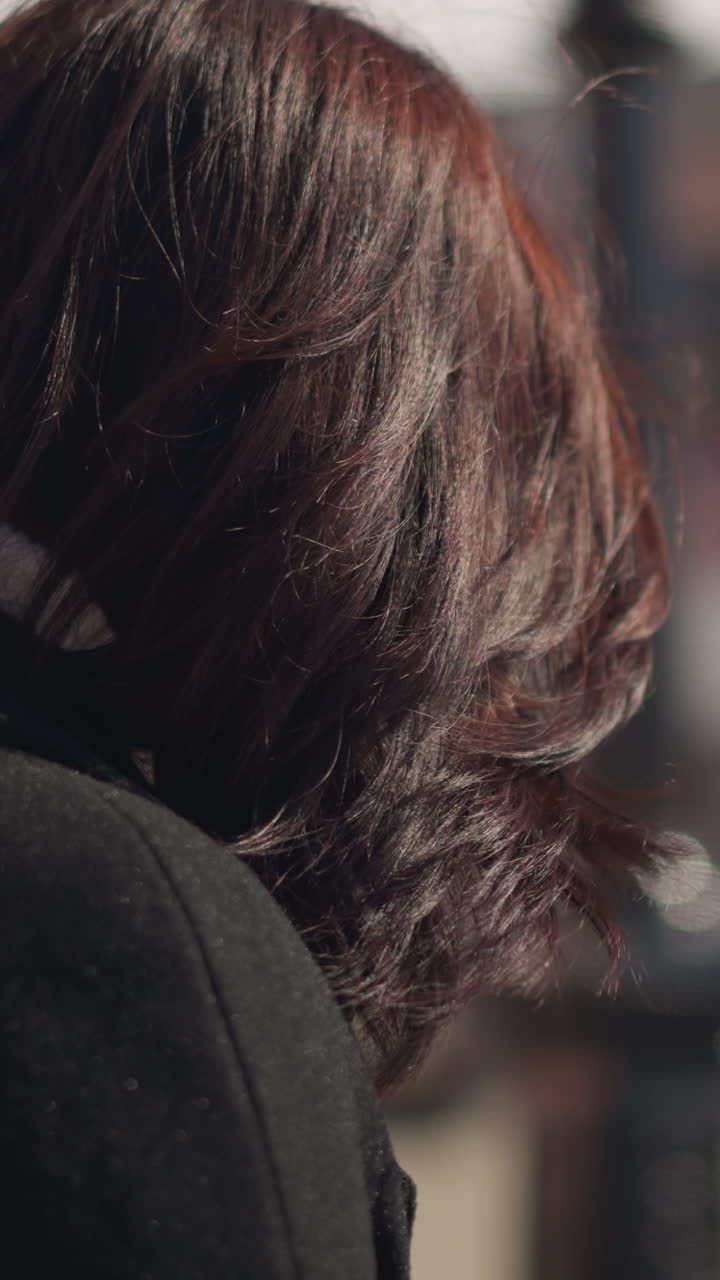 primer plano de una joven segura con un pequeño anillo en la nariz, mirando hacia abajo y luego girándose para mirar hacia adelante mientras la luz del sol destaca su cara, con farolas y grandes tiendas en el fondo