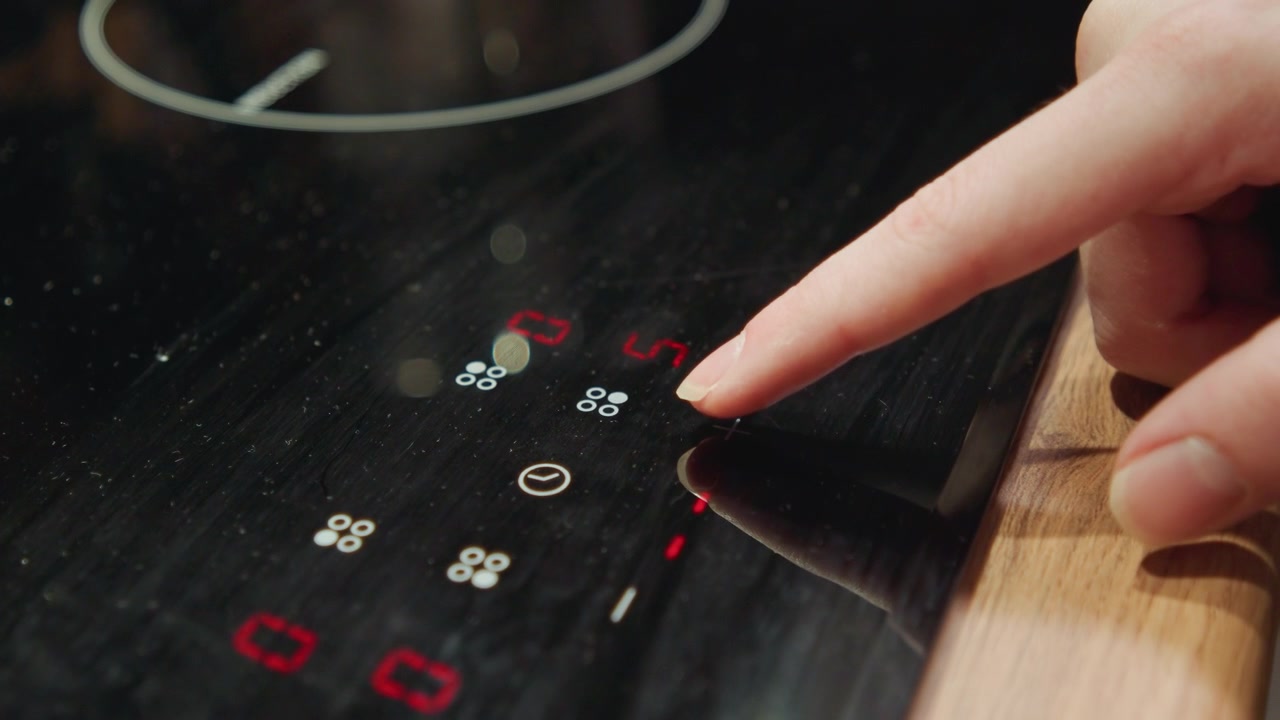 Tight overhead-right close-up showing hand fingertip pressing glowing touch control on glossy black induction cooktop, with illuminated symbols, red status light, glossy reflections, wooden