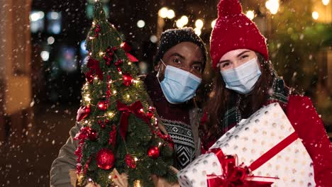 vista de cerca de una feliz pareja multiétnica usando máscaras faciales y sosteniendo regalos de navidad mientras sonríe a la cámara en la calle mientras nieva en navidad