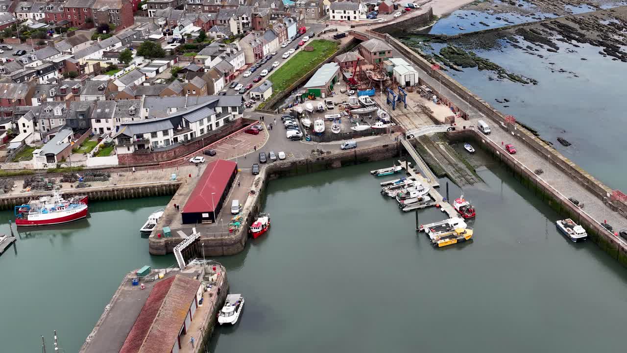 Drone glides above harbor, boats, and shoreline in a historic Scottish coastal town, daylight