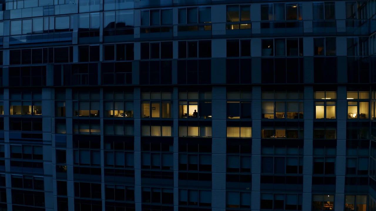 Un edificio per uffici in vetro scuro riempie l'inquadratura di notte, con luci calde sparse dalle finestre che formano un motivo geometrico sulla facciata esterna e creando un'atmosfera tranquilla