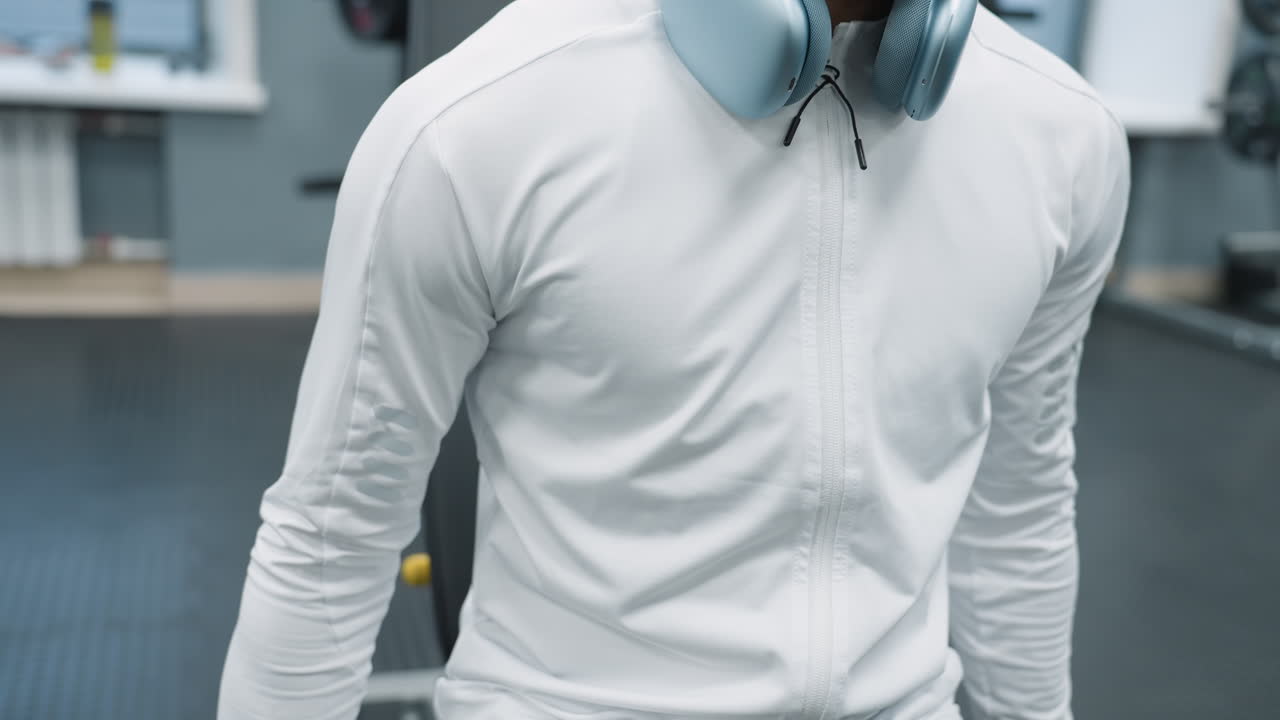 close up portrait of fitness coach in white jacket with headphones lifting dumbbell for muscle training inside modern gym, focused facial expression indicates determination during workout session