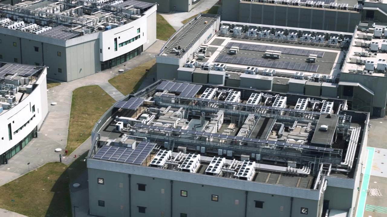 High-angle aerial shot flying over a large, modern data center complex featuring multiple buildings with solar panels and cooling systems on the roofs