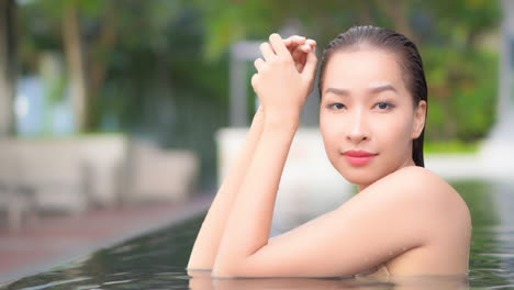 beautiful asian woman with wet hair bathing in a pool looking at camera while leaning elbows on the edge of the swimming pool