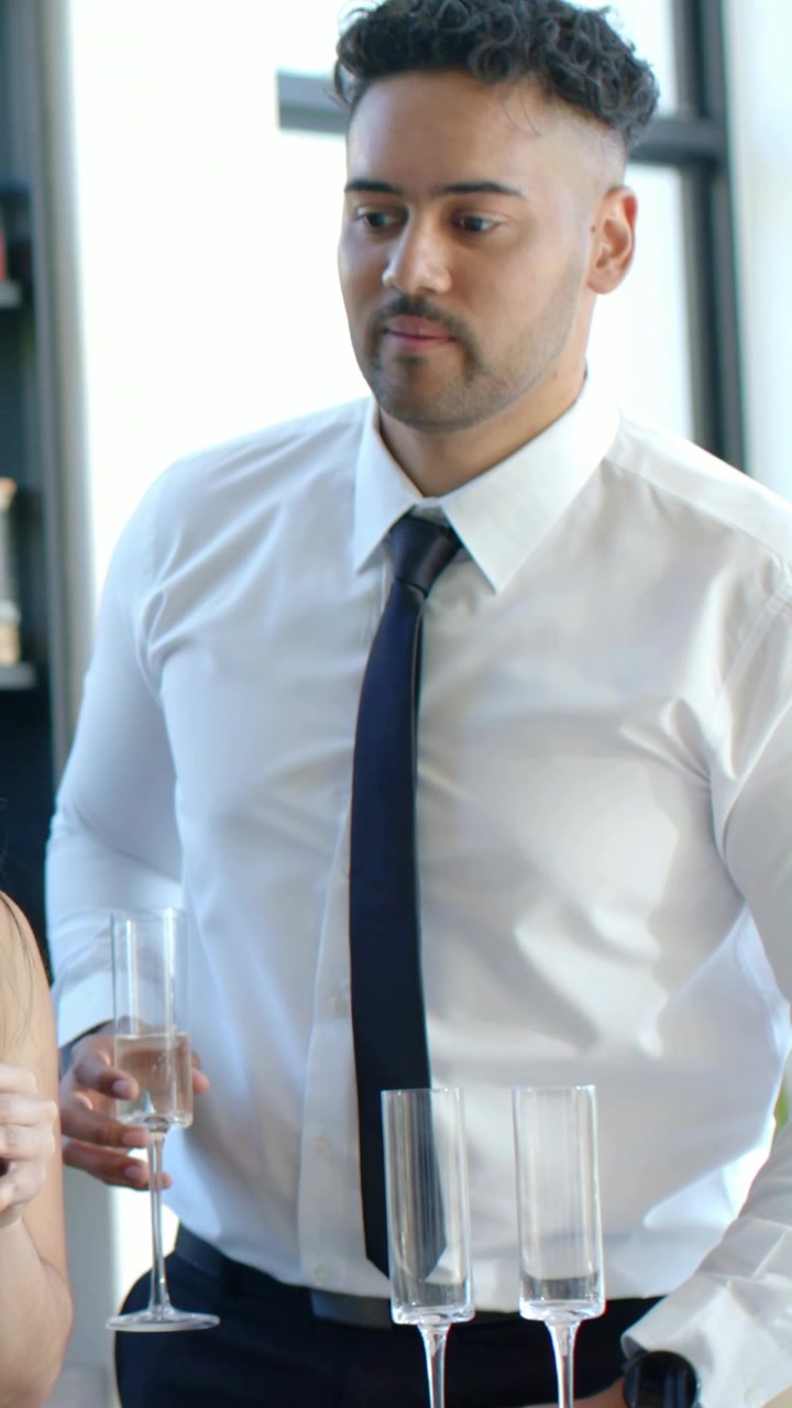 Video verticale: Uomo in camicia e cravatta blu navy che si allunga per sistemare bicchieri e piatti in cucina, servendo una bevanda.