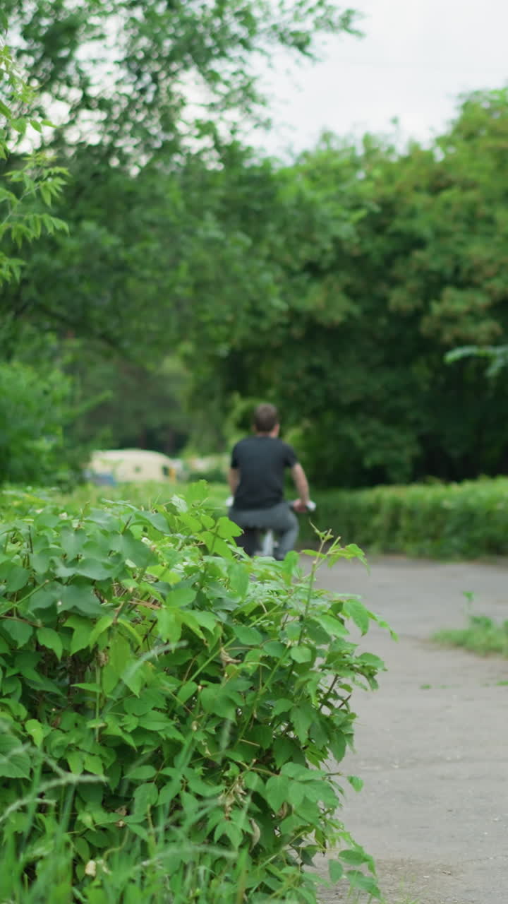 vista trasera de un niño con pantalones grises y camiseta negra montando su bicicleta a lo largo de una carretera pavimentada rodeada de vegetación exuberante y árboles, con una vista borrosa de coches moviéndose en el fondo