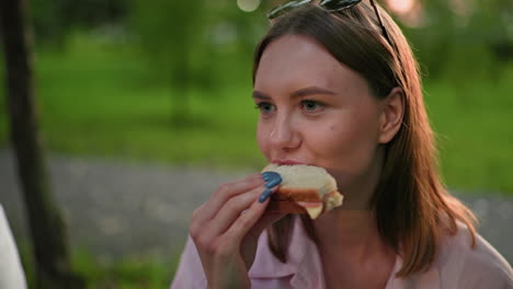 dama de top rosa con gafas de sol descansando en la cabeza comiendo sándwich a la parrilla mientras está sentada al aire libre, con vista parcial de alguien cerca en top blanco y fondo verde borroso con árboles