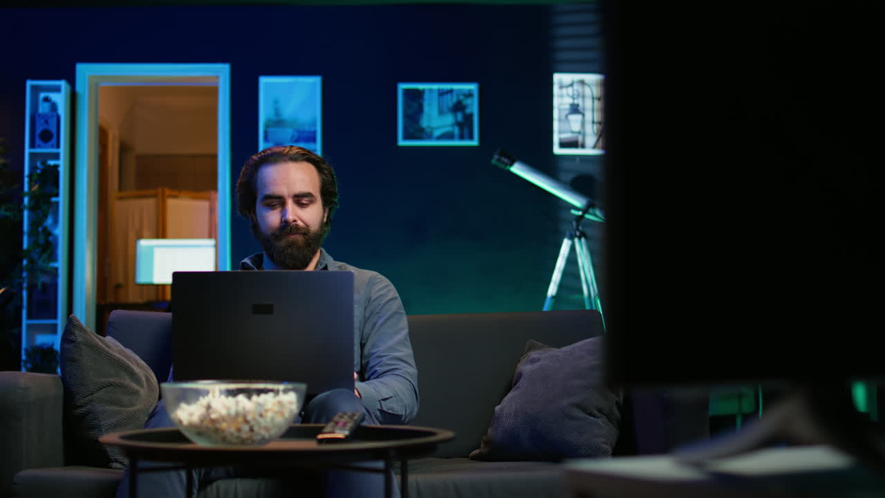 Freelancer working on laptop while watching TV in apartment office