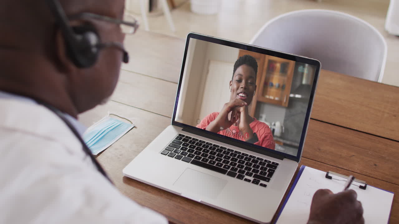 Afroamerikansk forretningsmand sidder ved skrivebord, bruger bærbar computer og har videoopkald med en kvindelig kollega.