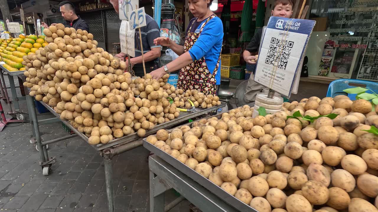 Vendors at an outdoor market in Kanchanaburi, Thailand, arrange longkong and oranges on carts. Daylight, handheld camera, bustling street atmosphere