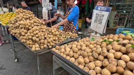 vendors at an outdoor market in kanchanaburi, thailand, arrange longkong and oranges on carts. daylight, handheld camera, bustling street atmosphere