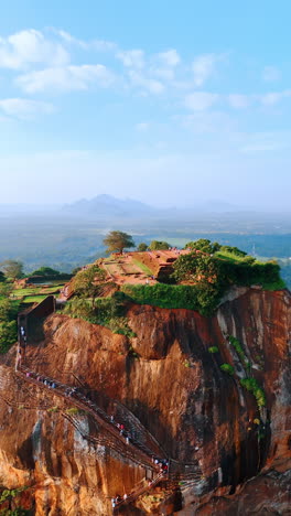 distancing from a brown lava-originated rock with the stairs for tourists climbing to the top. aerial perspective on sigiriya in sri lanka at the backdrop of hazy landscape and blue sky. vertical video.