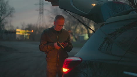 a man in a green jacket stands beside an open car trunk, holding a handgun and a phone, with dim light seen around