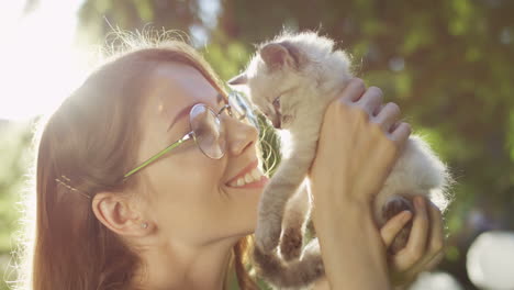 close-up view of a caucasian woman in glasses holding small cat and kissing it in the park on a summer day