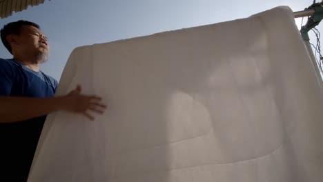 an asian man takes down bed sheet that were drying on a clothing rack on his apartment balcony in the bright sunshine