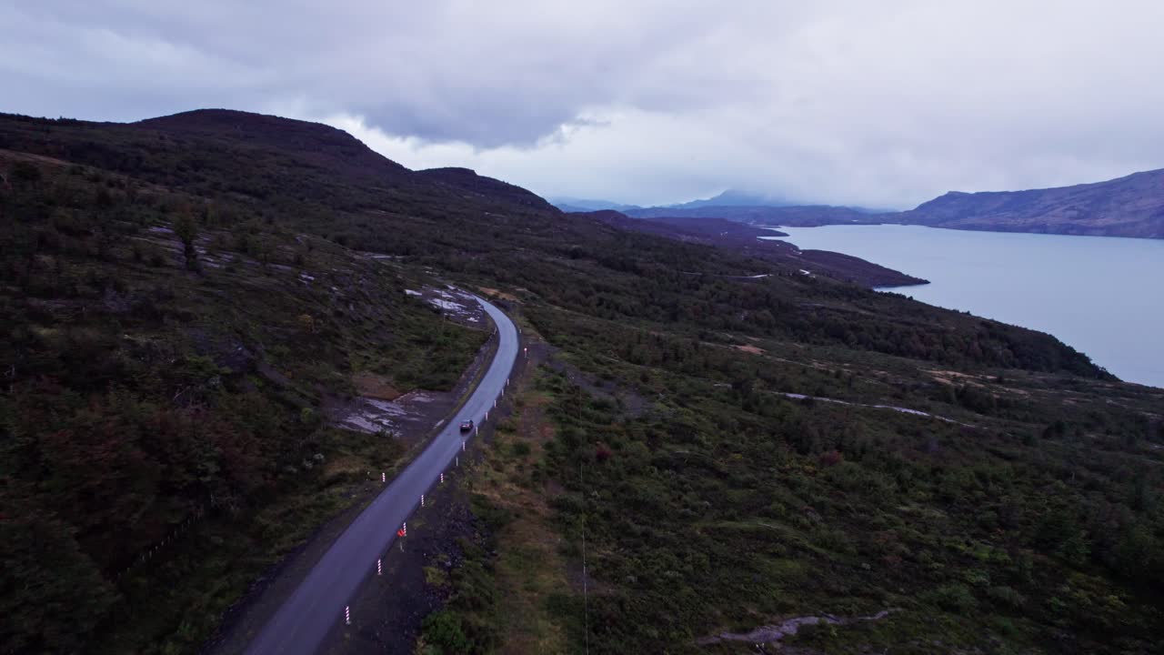 A road winding through the desolate, dark green landscape of Torres del Paine, Chile. Features a large, cold-toned lake fjord to the side, under a cloudy, dramatic sky, emphasizing isolation
