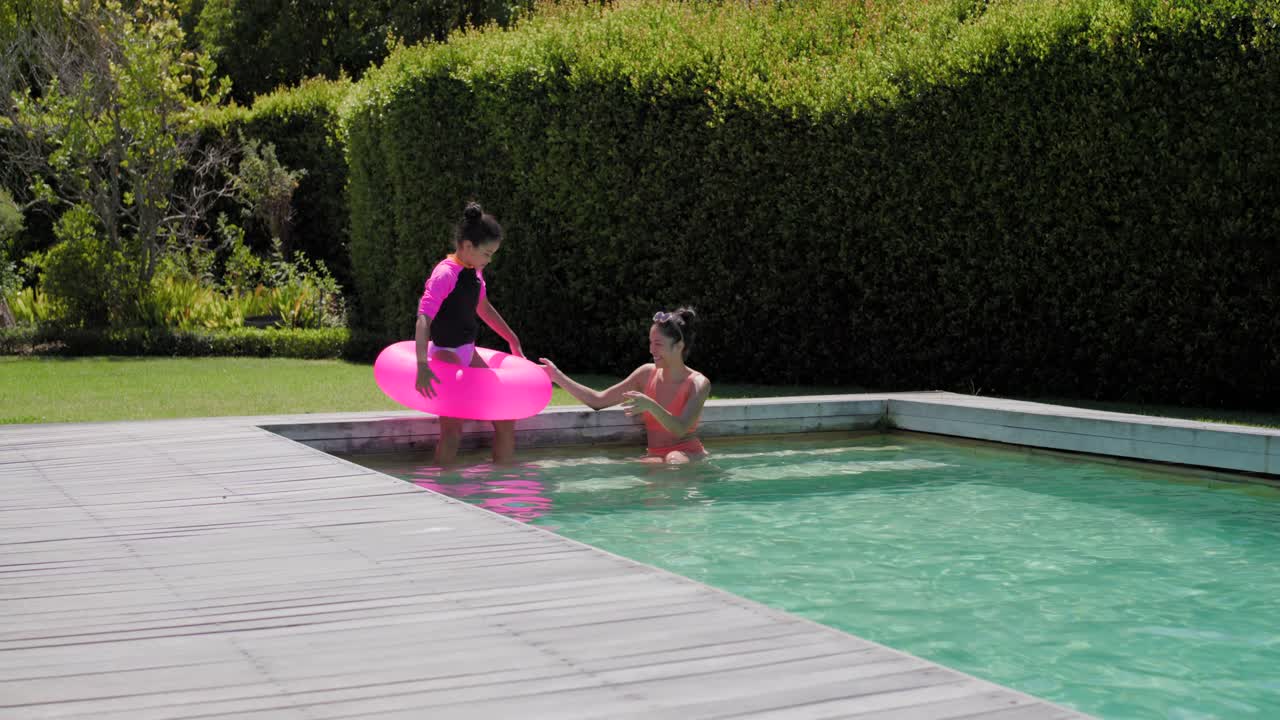 African American woman and girl holding pink ring, stepping into backyard pool, learning to float