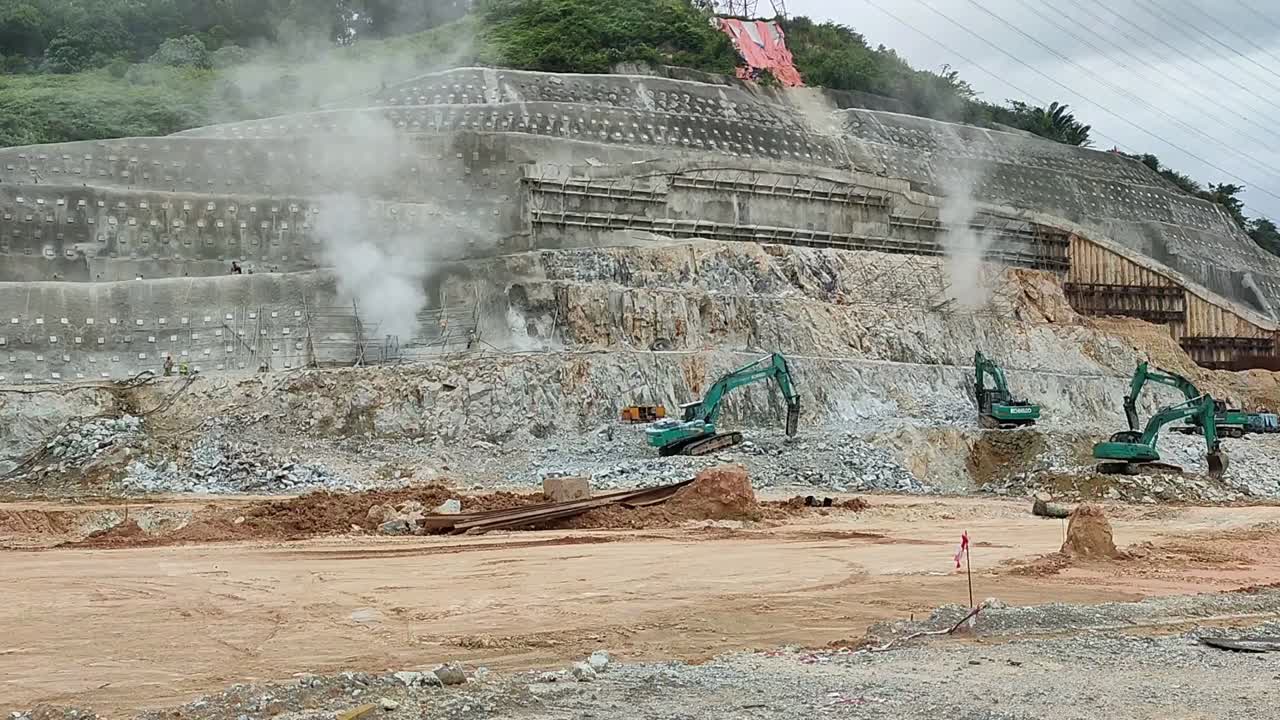 Heavy machinery, including excavators and trucks, operates on-site for earthwork. Rocks are broken, and soil is leveled. Excess debris is cleared, ensuring progress in the construction area.