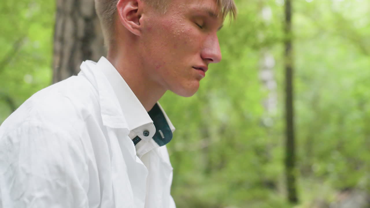 Close side view botany student in white shirt putting on glasses in forest with tired expression surrounded by blurred trees, reflecting fatigue and exhaustion during outdoor work in natural setting