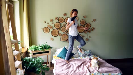 playful young lady is singing in blow dryer, dancing and jumping on bed and listening to music while pet dog is lying on bed in nice modern style apartment.