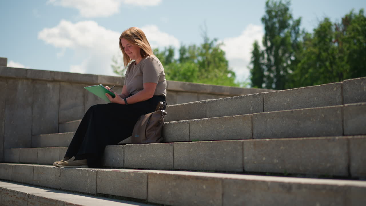 lady sitting alone on stone steps writing on clipboard under warm sunlight, wearing beige top and black skirt, focused and calm expression, handbag beside, surrounded by open air and greenery
