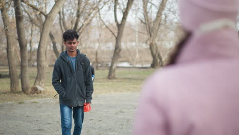 Young man with dark curly hair wearing gray hoodie and jeans standing outdoors in park holding orange gloves, carrying black backpack with water bottle, looking down with serious expression early spring day