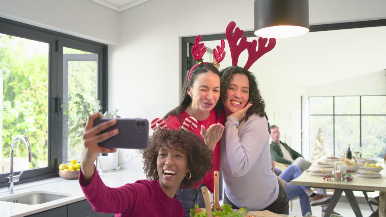 Diverse female friends making salad at kitchen island showing smartphone for selfie wearing antlers