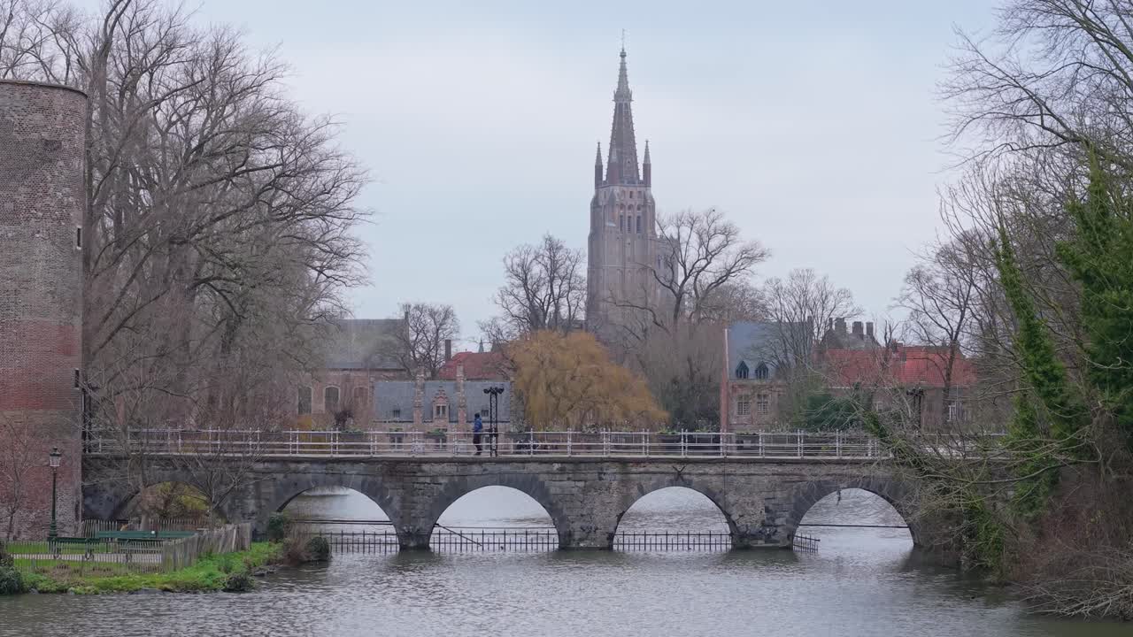 Inquadratura statica del Minnewaterbrug (Ponte degli Innamorati) con un passante, che mostra il Campanile della Chiesa di Nostra Signora nel centro storico di Bruges.