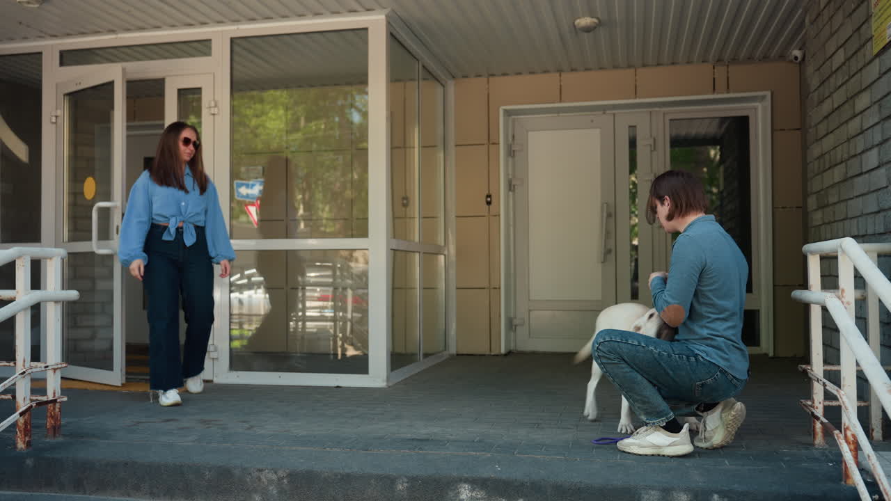 아파트 입구에서 래브라도를 데리고 있는 백인 여성과 남성, 사진작가가 무릎을 꿇고 솔직한 재회 장면을 촬영하고 있어요. 유리문, 금속 난간, 콘크리트 계단, 화창한 정오, 캐주얼한 청바지와 스니커즈, 즐거워하는 반려동물이 보이네요.