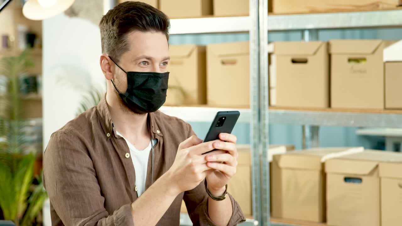 Cucasian designer man wearing black facial mask and texting on smartphone in a fashion clothing store