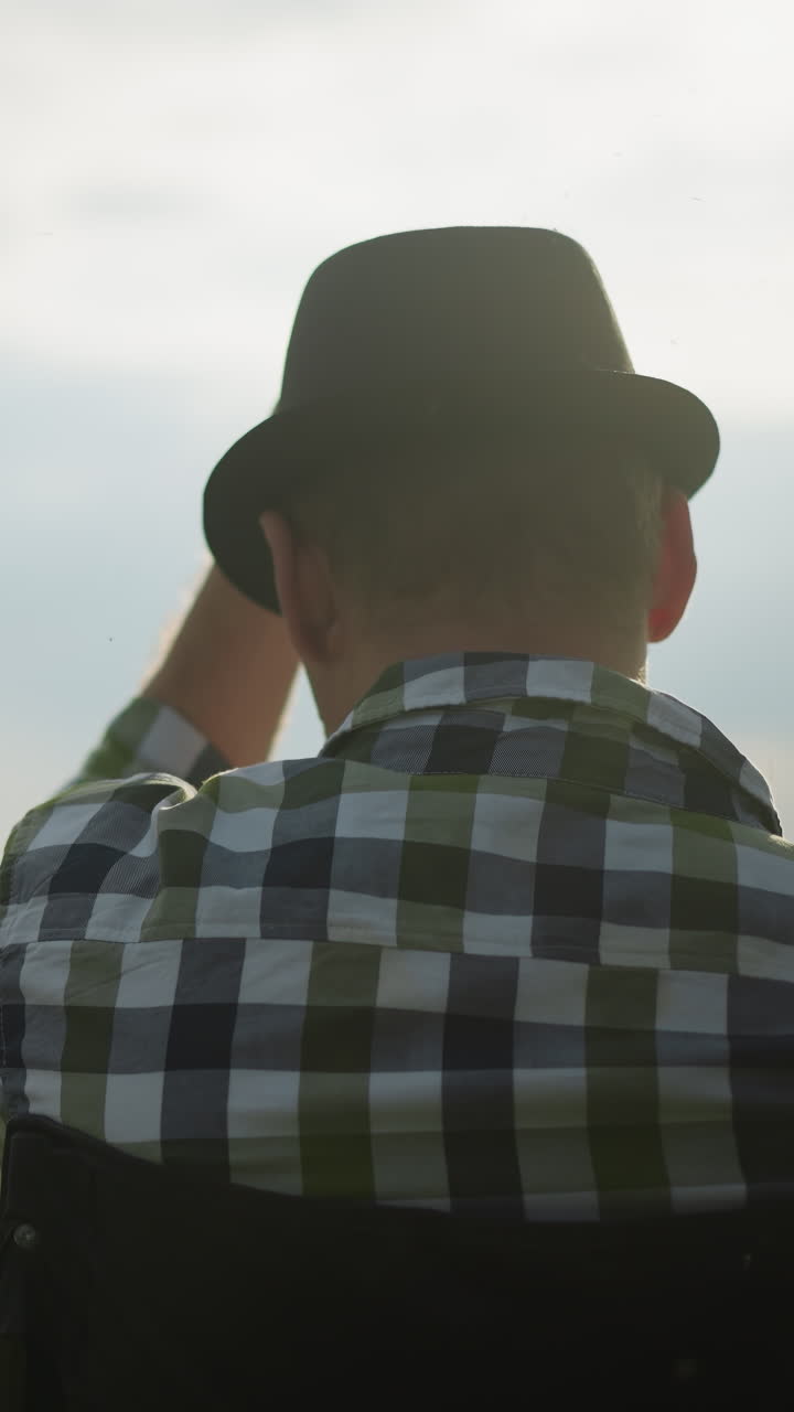 A back view of a man wearing a black hat, adjusting it as he sits in a grassy field. In the background, a blurred figure of a woman is painting, creating a outdoor artistic scene under a cloudy sky