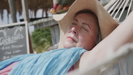 relaxed senior caucasian woman lying on hammock sleeping in the sun by beach bar, in slow motion