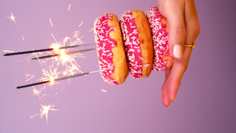 Woman holding Birthday Donuts with burning sparkler as candles in her Hand. Pink lighted donut Cake for celebration with colorful light purple background. Happy Birthday or anniversary concept.