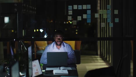 video of tired african american businessman at desk using laptop, working alone at night in office