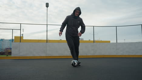 hombre controlando hábilmente la pelota de fútbol con los pies en la arena deportiva durante la sesión de entrenamiento, el fondo presenta edificios urbanos, esgrima y postes de portería