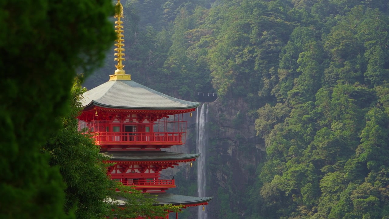 Ripresa suggestiva del tempio Seiganto-ji che emerge dal verde in primo piano con la cascata di Nachi che scende attraverso il sacro paesaggio montano di Kumano
